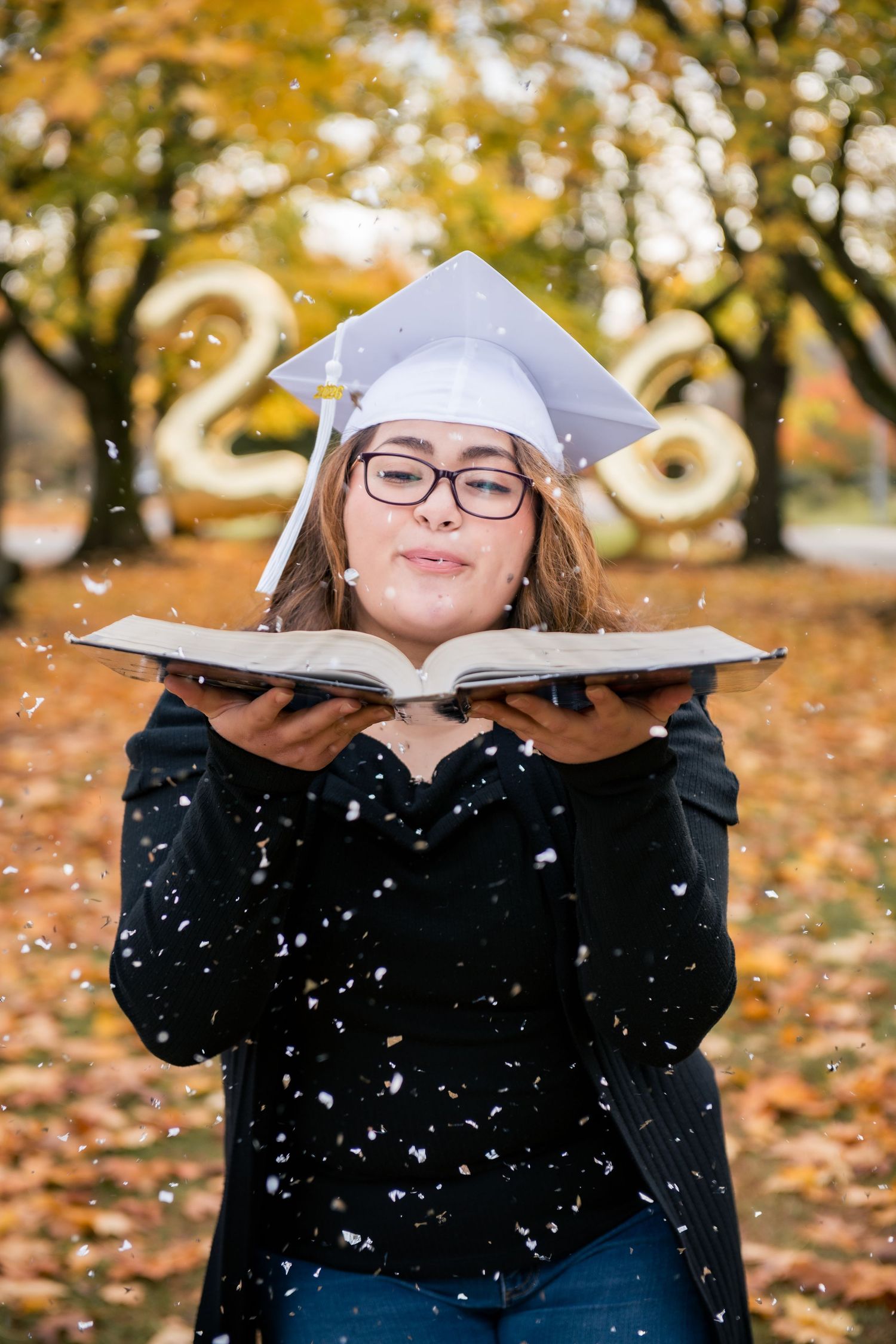 A person in a graduation cap and black sweater holds open a book while autumn leaves and snow fall in the background.