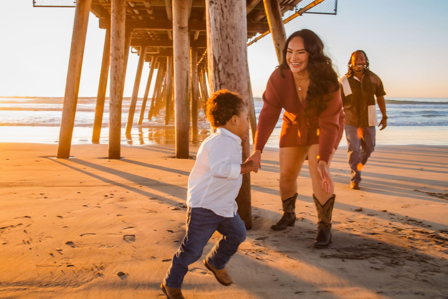 People walking along a sandy beach at sunset with long shadows cast by a wooden pier overhead.