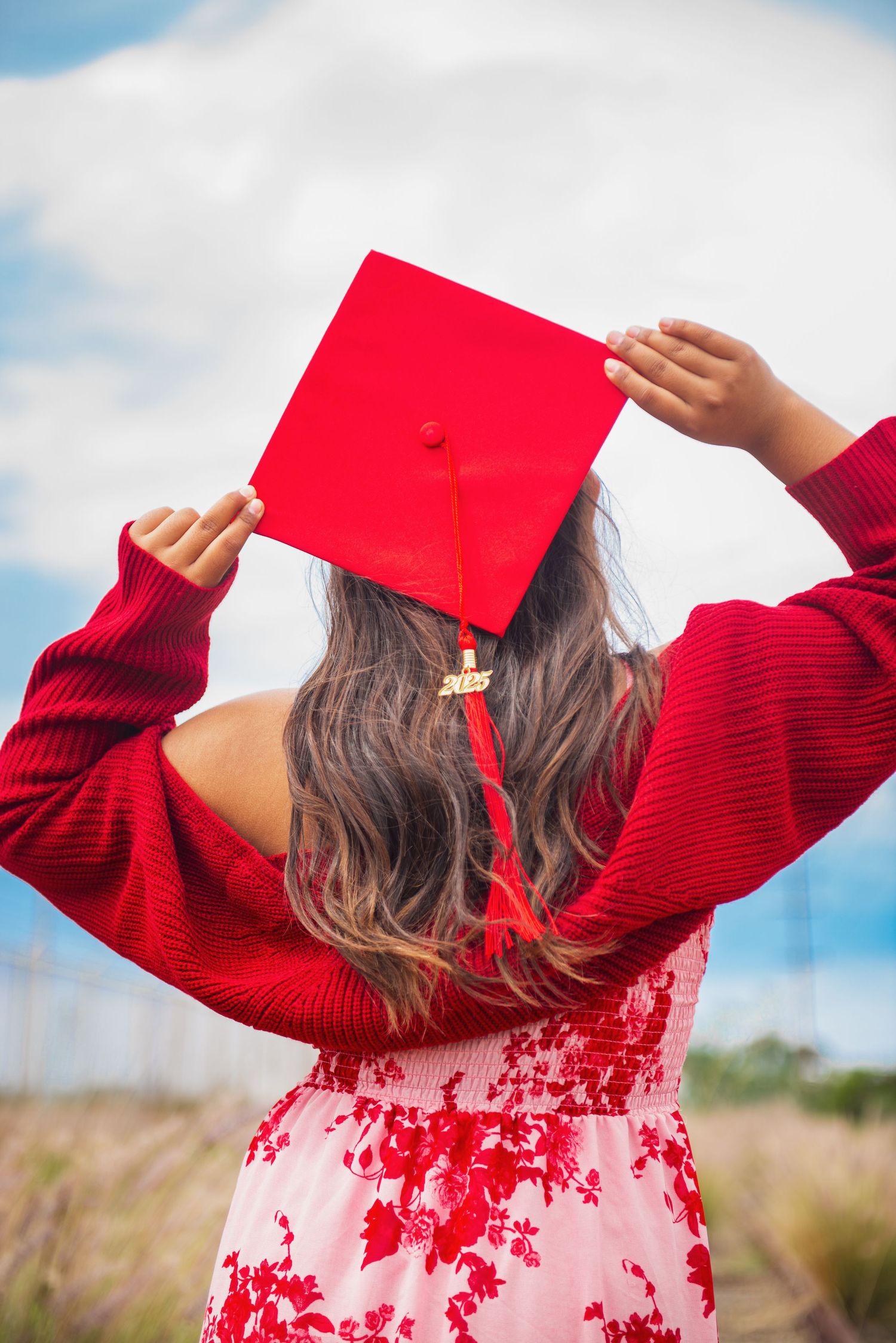 Graduate in red dress and sweater holds up graduation cap against blue sky and desert landscape in celebration.