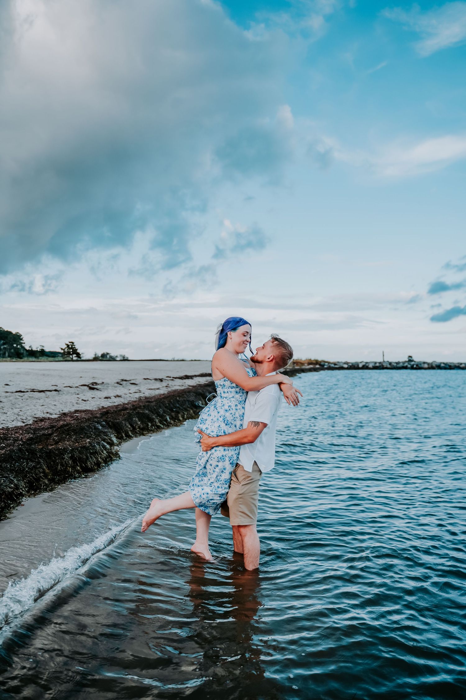 A couple shares a romantic moment by the ocean at sunset with a stone jetty in the background.