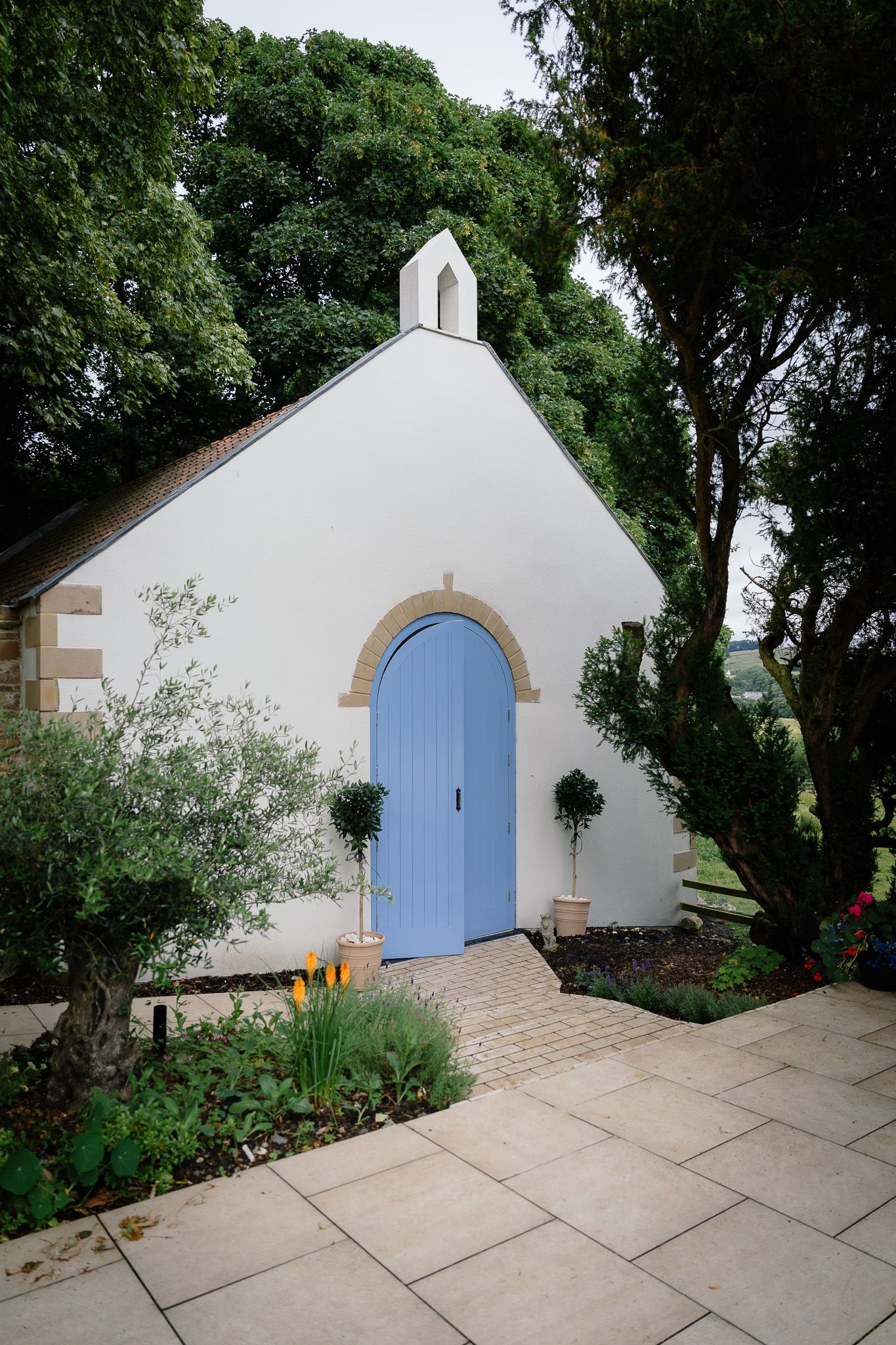 White chapel building with blue door and stone pathway surrounded by lush greenery. Woodhill Hall.