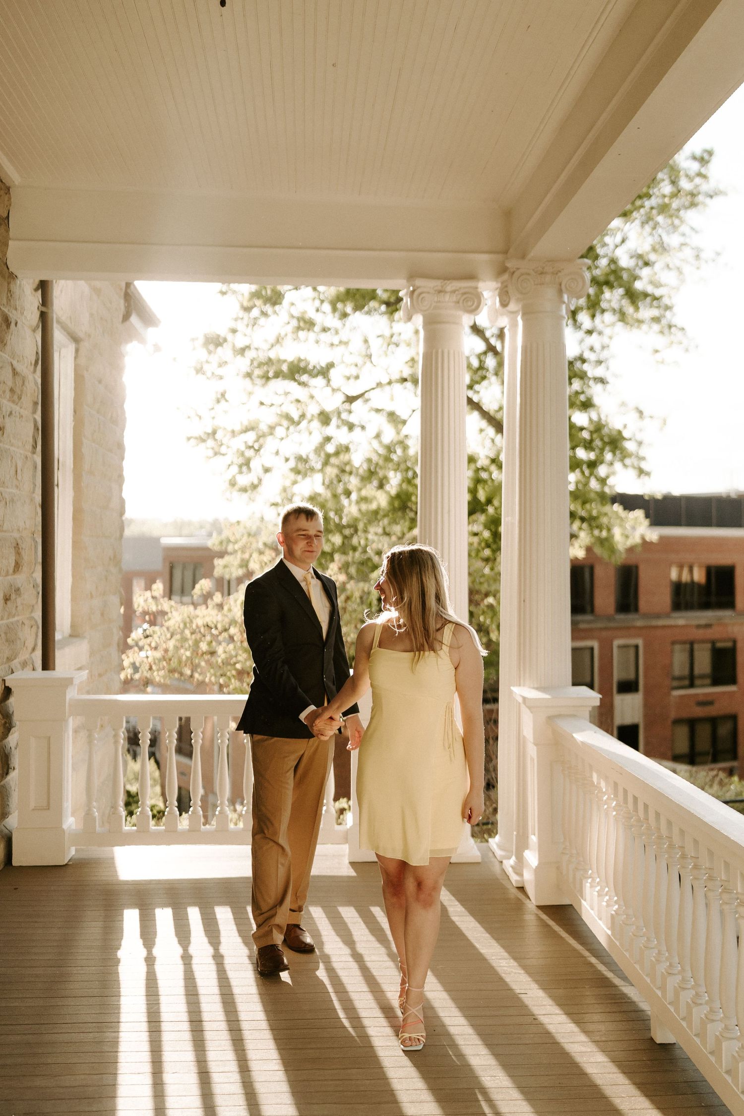 Couple holds hands walking on sunlit historic porch with white columns.