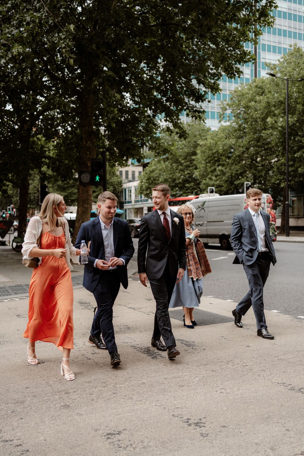 A group of wedding guests walking down a tree-lined city street, dressed in formal attire.