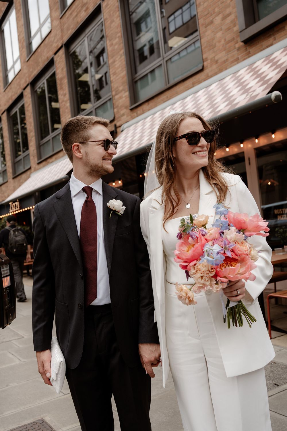 A couple in wedding attire walks together on a city street with a colorful floral bouquet.