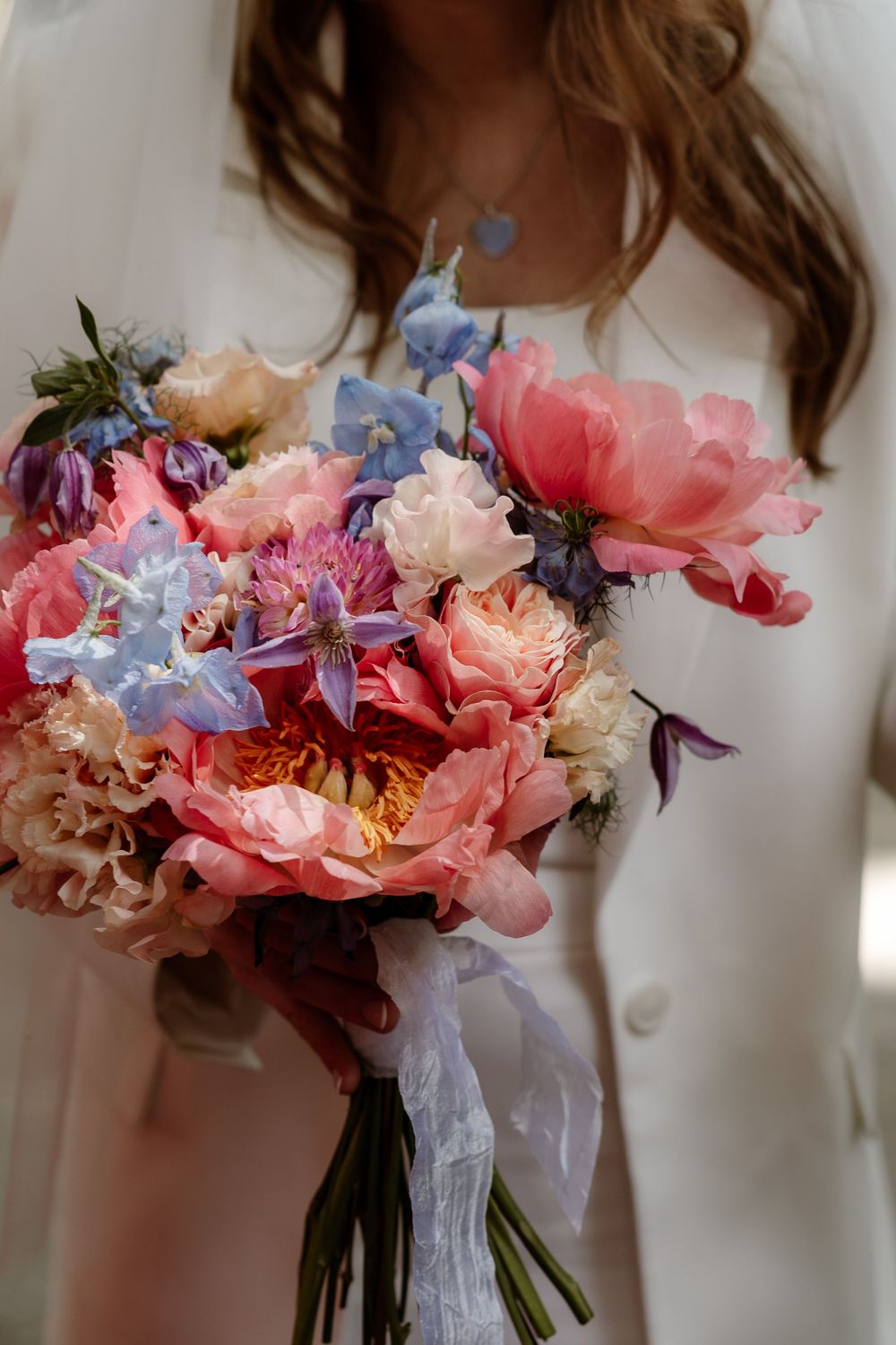 A romantic bridal bouquet featuring pink peonies, peach roses, and delicate blue delphiniums tied with white ribbon.