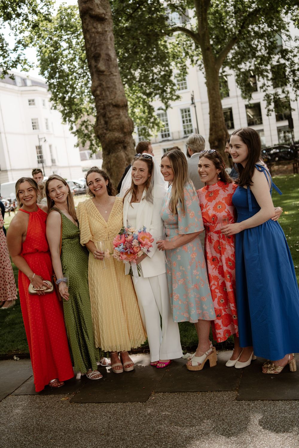 A group in colorful dresses and outfits pose together outdoors in front of historic white buildings on a sunny day.
