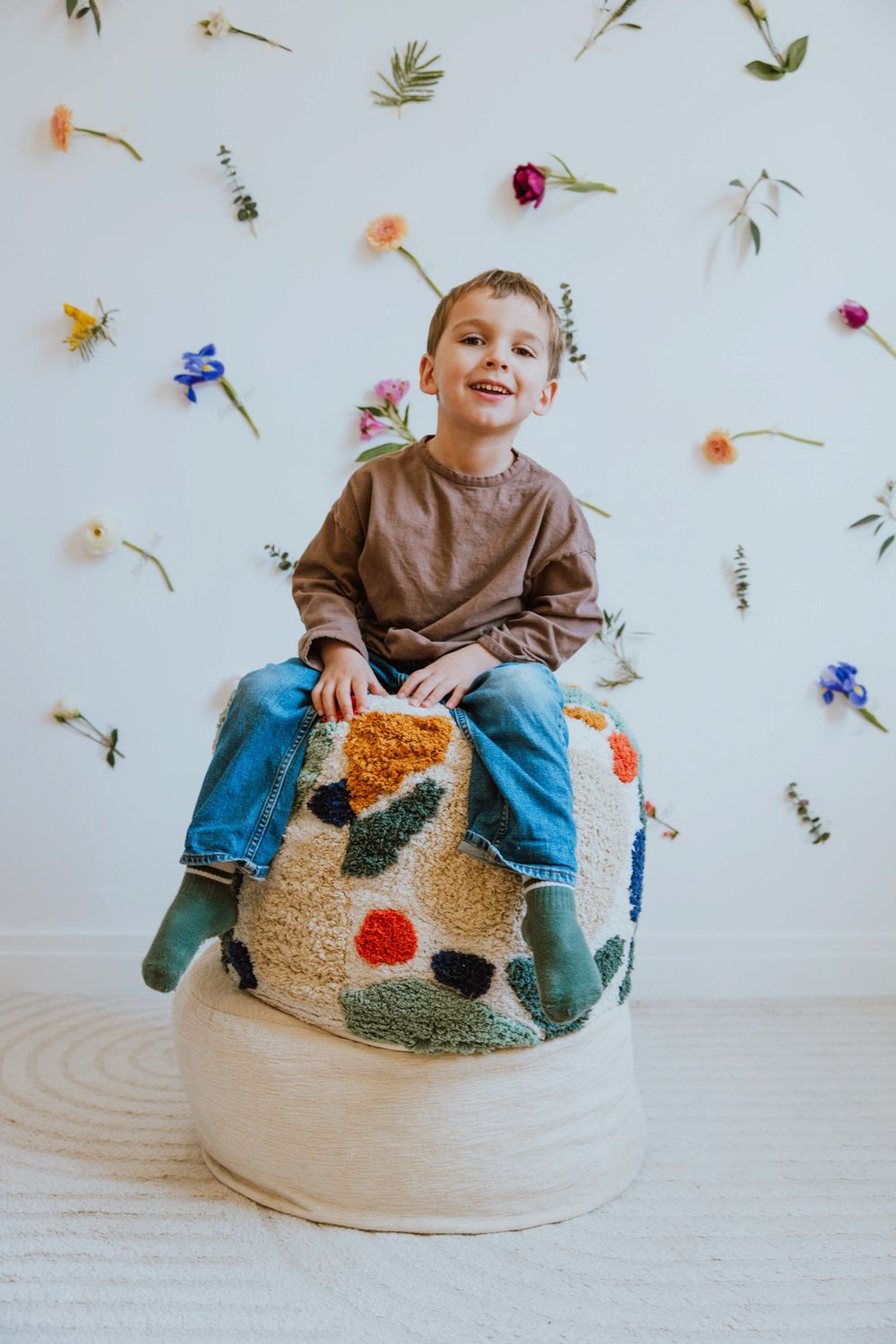 A child sits on a round beanbag chair against a white wall decorated with scattered pressed flowers and botanicals.