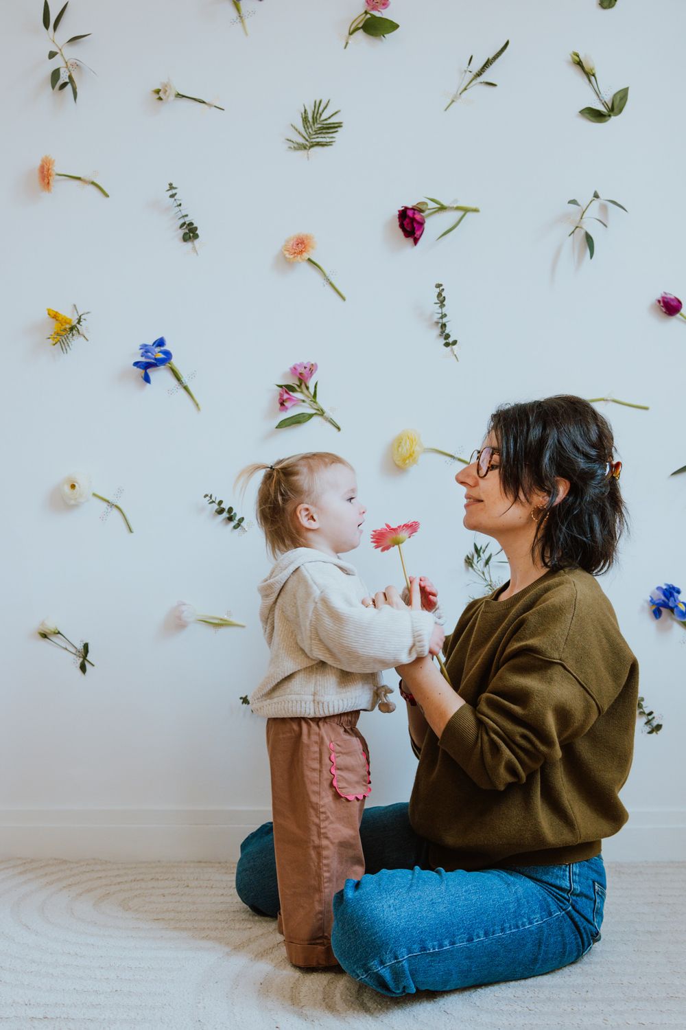 Two people sit on floor playing with falling flower petals against white wall in artistic photo series.
