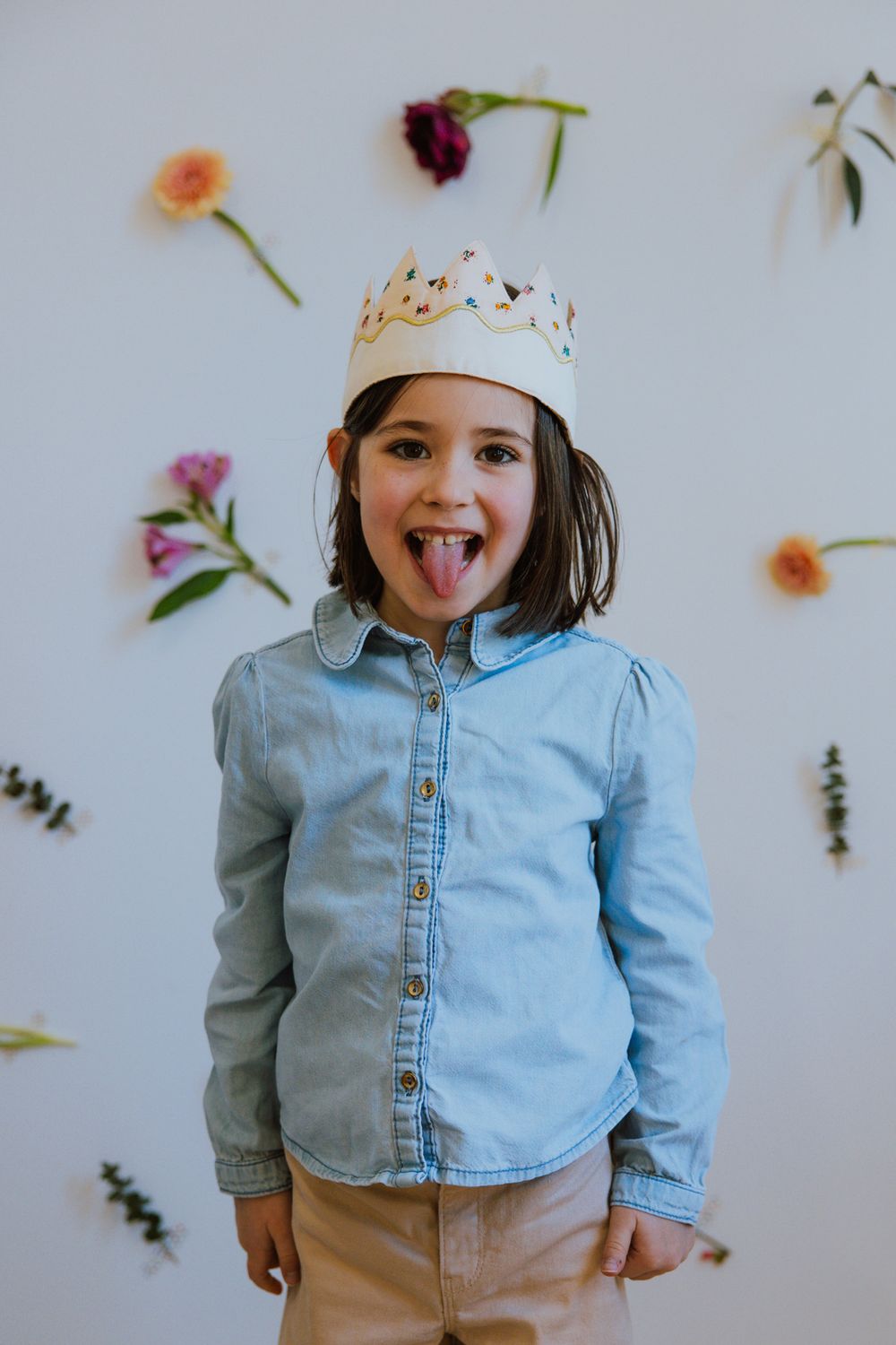 Child wearing paper crown and denim shirt poses playfully against white background with scattered flower petals.