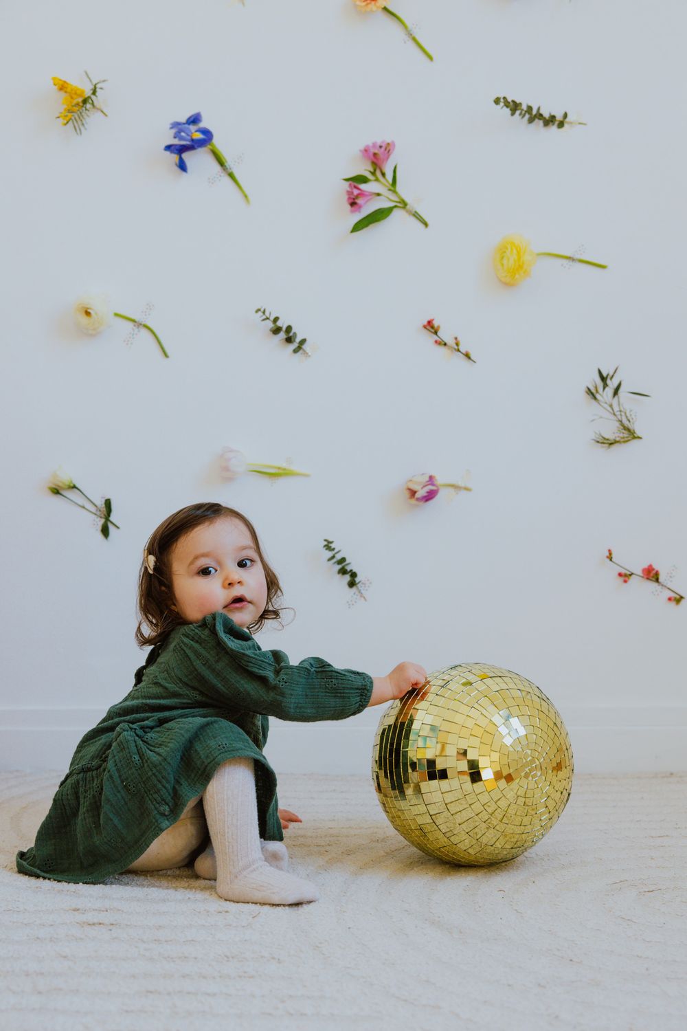 A small child in a green jacket plays with a shiny gold disco ball on a white floor with scattered flower petals.