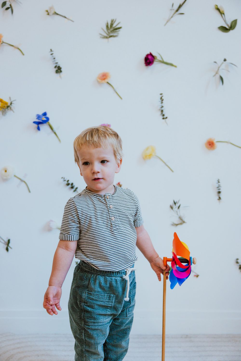 A toddler in denim overalls and striped shirt stands against a white wall decorated with scattered flowers and petals.