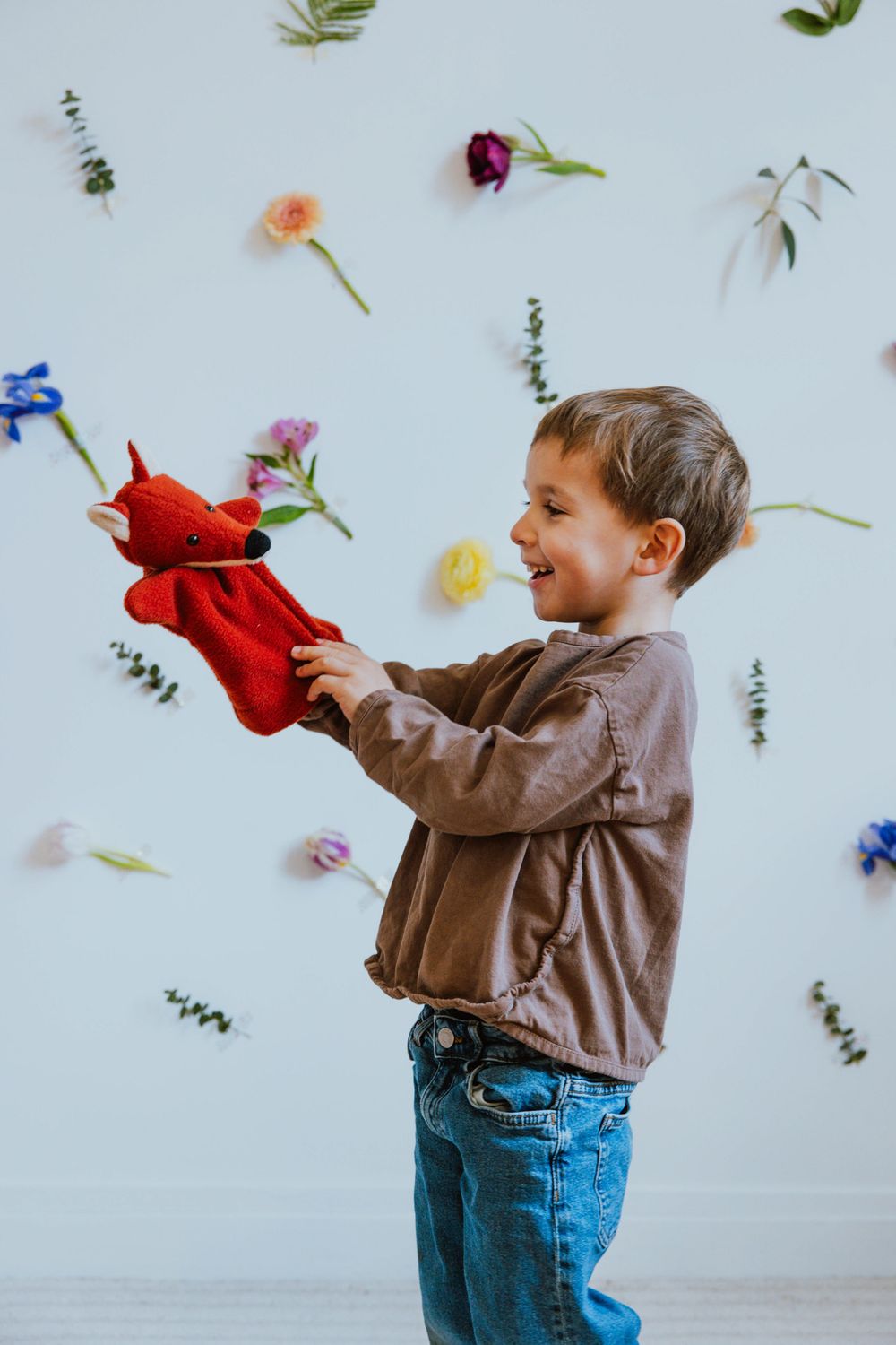 Child playing with red puppet against white wall decorated with scattered colorful flowers.