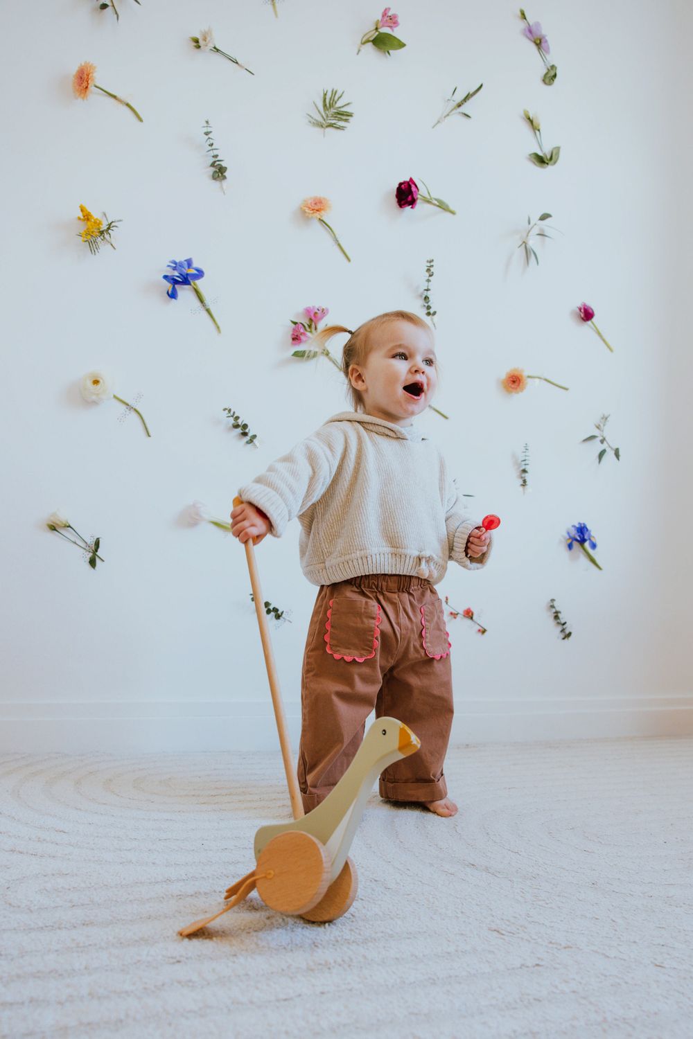 A joyful toddler plays with a wooden duck pull toy against a white wall decorated with scattered colorful flowers.