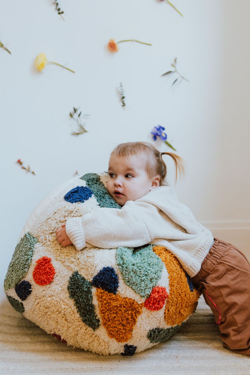 Small child leans against large colorful plush pouf ottoman with floral wall decals in background.