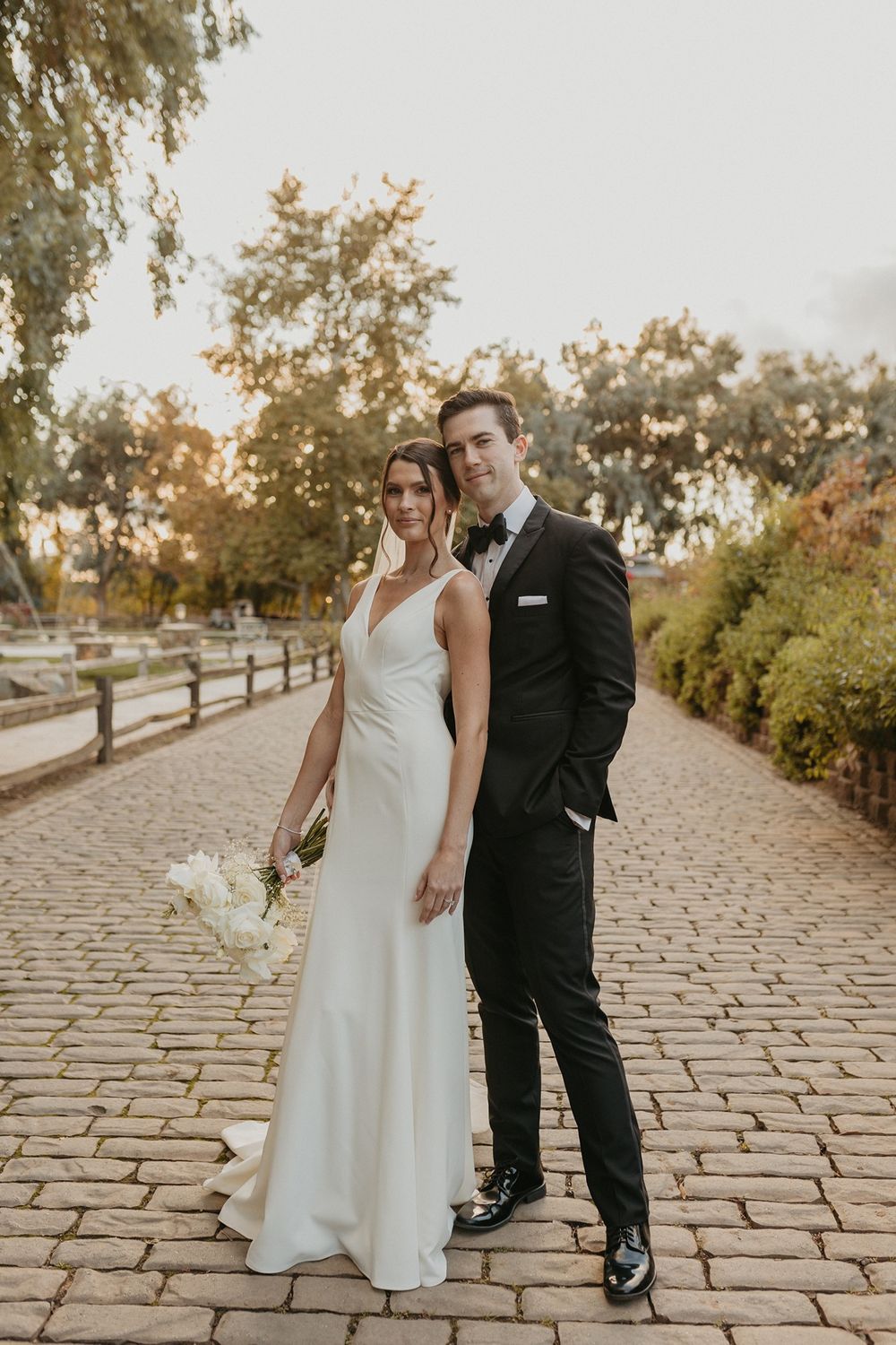 Elegant couple in wedding attire pose on cobblestone path with trees and golden sunset light in background at Lake Oak Meadows in Temecula.