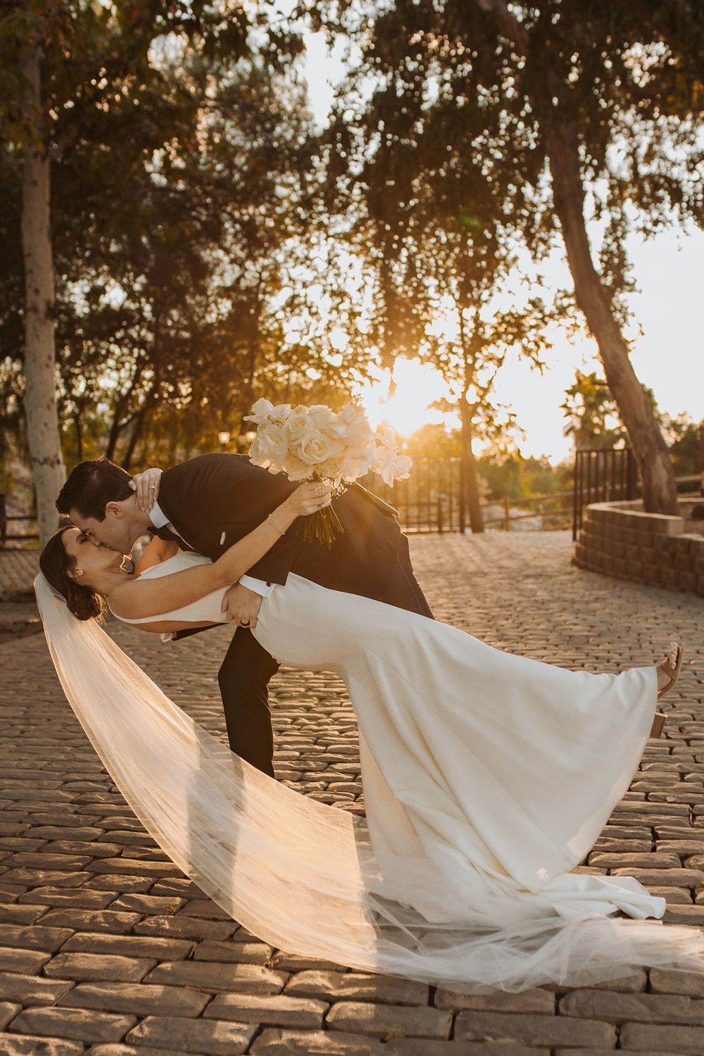 Romantic wedding dip pose on cobblestone path at sunset with flowing white dress and dramatic lighting at Lake Oak Meadows in Temecula.