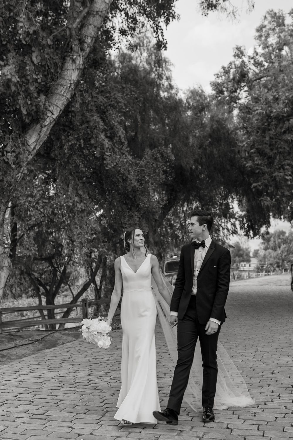A couple walks along a stone path surrounded by trees in black and white photography at Lake Oak Meadows in Temecula.