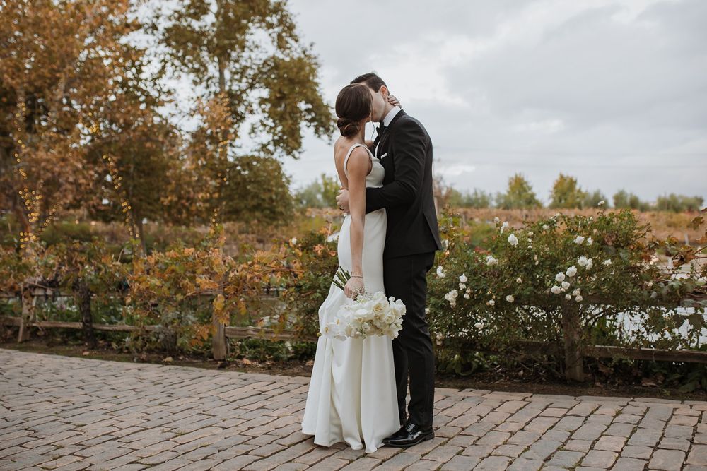 A romantic autumn wedding couple embraces on a stone path surrounded by fall foliage and changing leaves at Lake Oak Meadows in Temecula.
