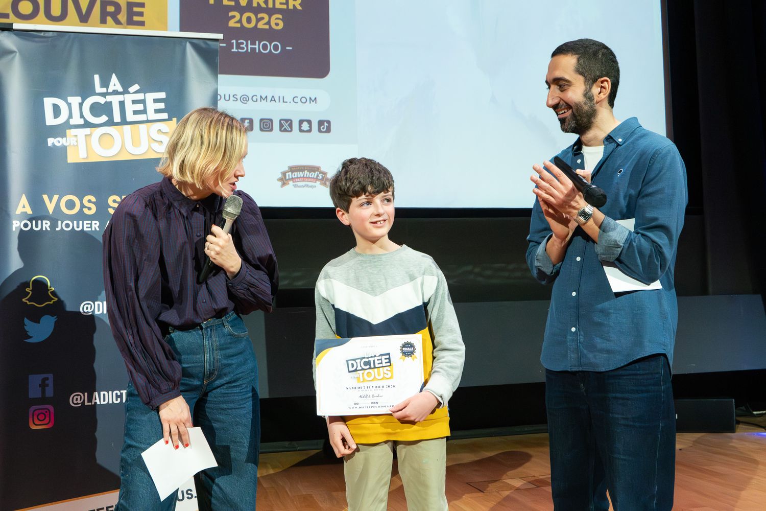 Students participate in a spelling competition event with judges and presenters on stage.