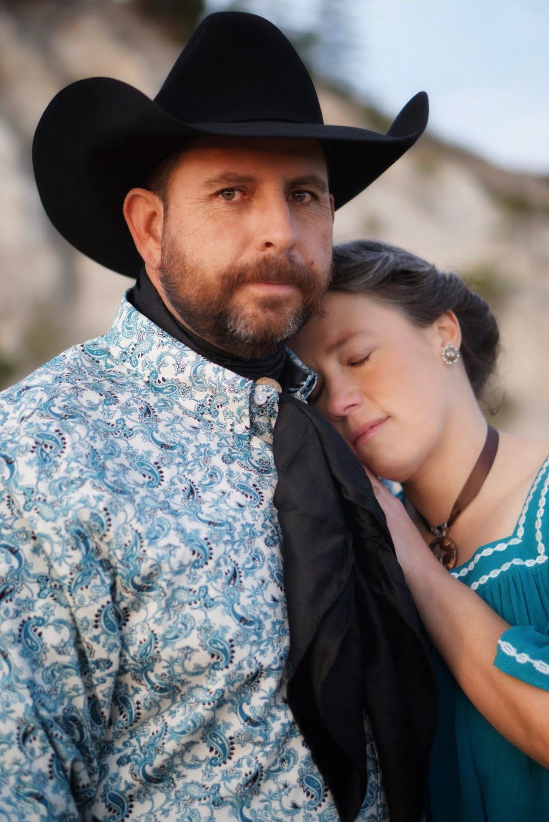 A couple in western attire share an intimate moment outdoors, with one wearing a cowboy hat and the other in a floral print.