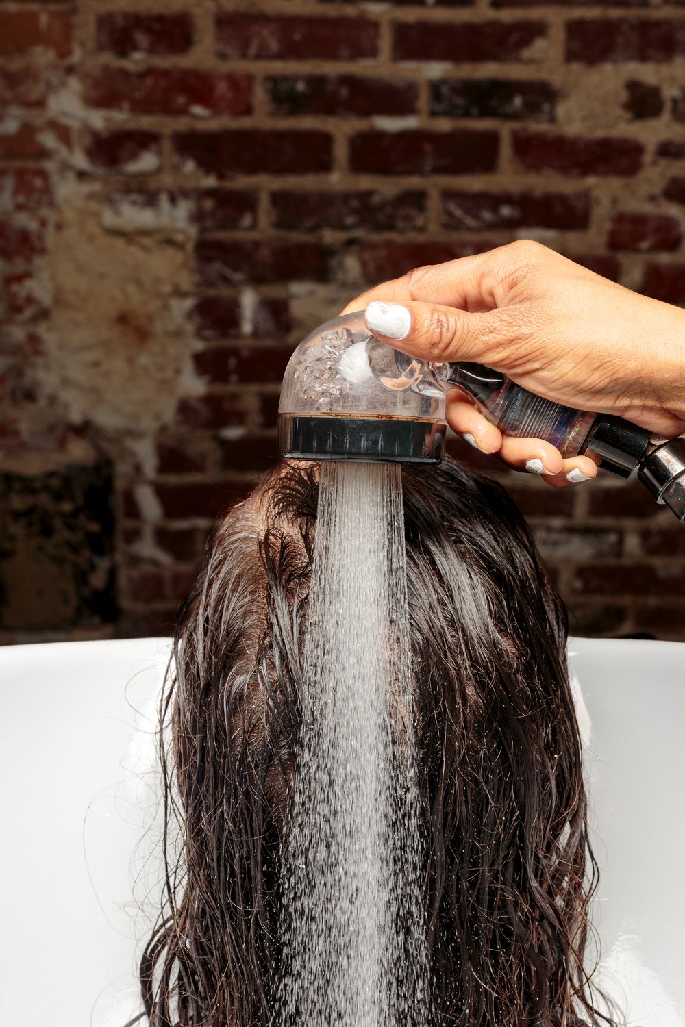 A hairstylist rinses long dark wet hair over a salon washing basin with a handheld shower head.