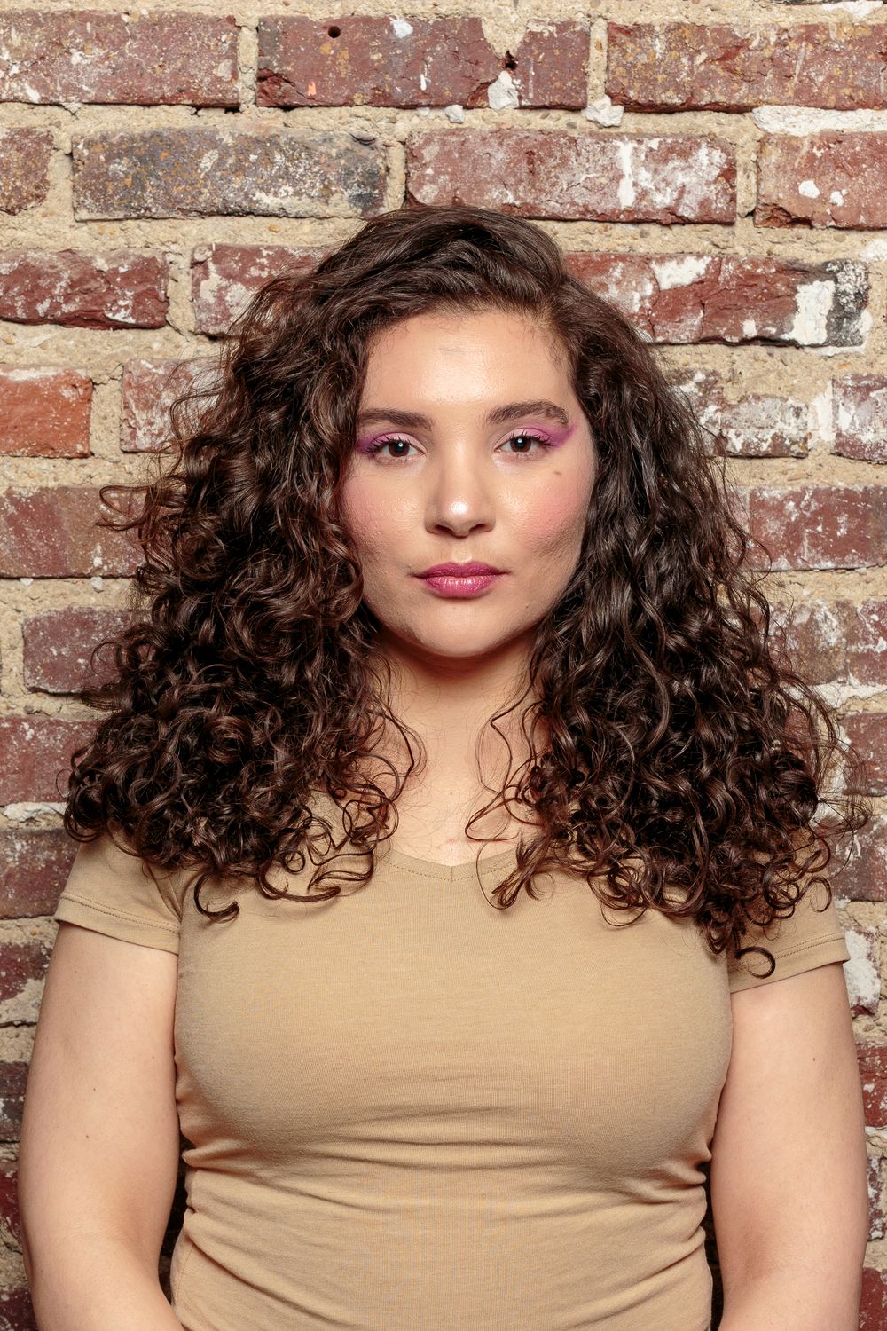 A young woman with long curly brown hair and pink eye makeup stands against a brick wall.