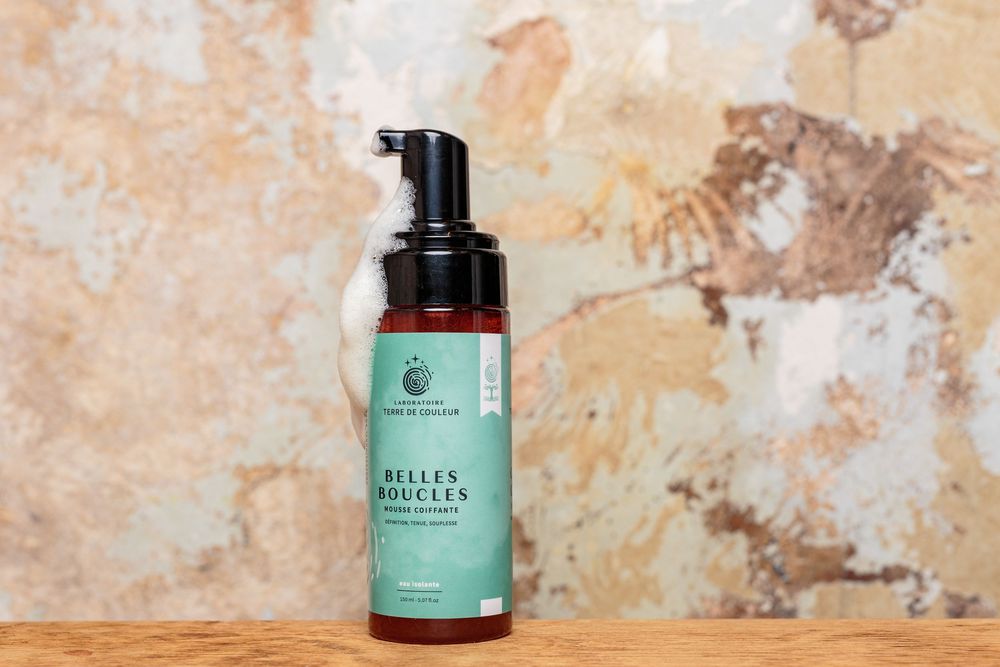 Belles Boucles curl foam product bottle displayed against rustic textured wall background.