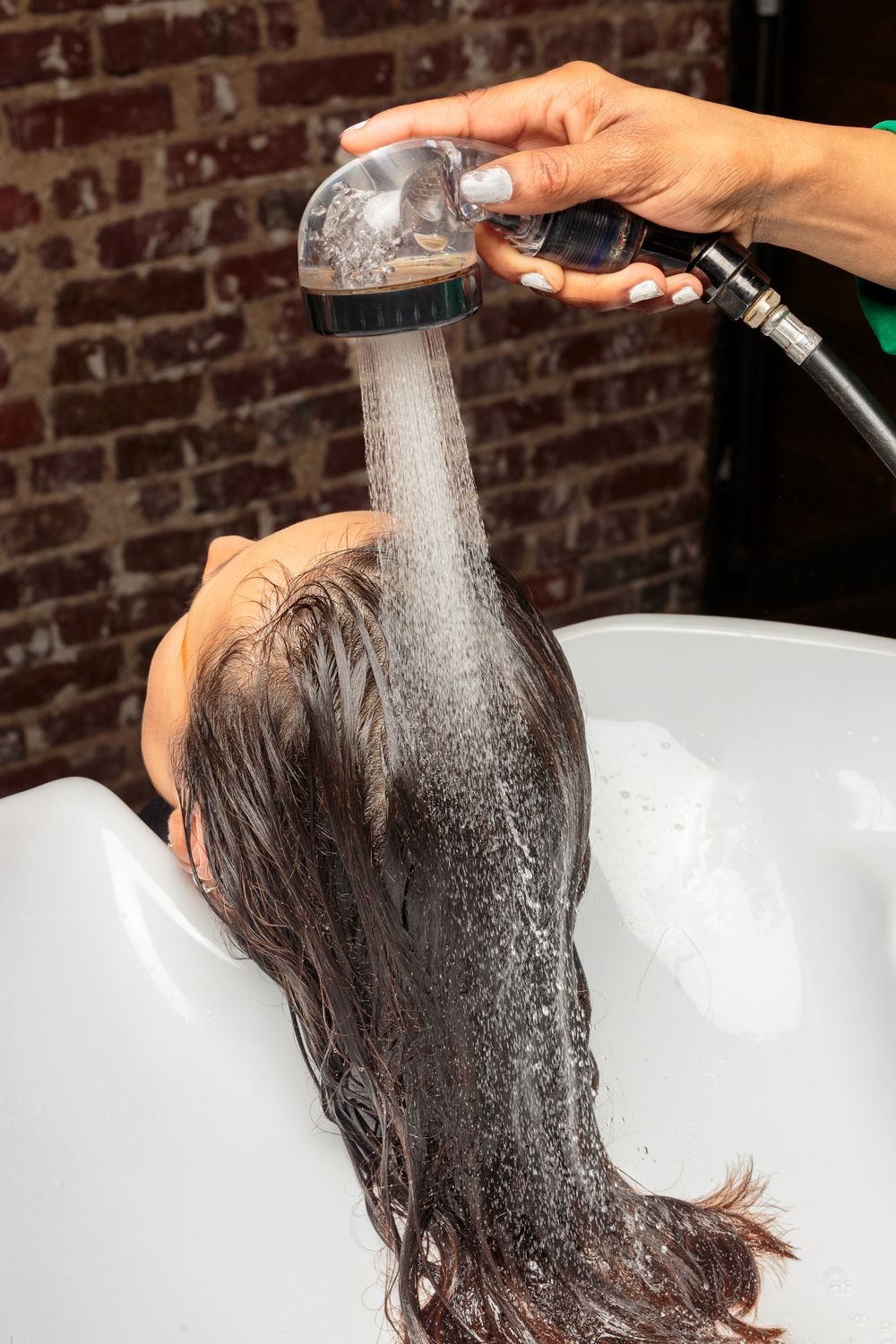 Professional hairstylist rinsing dark hair with a handheld shower at a salon wash basin.