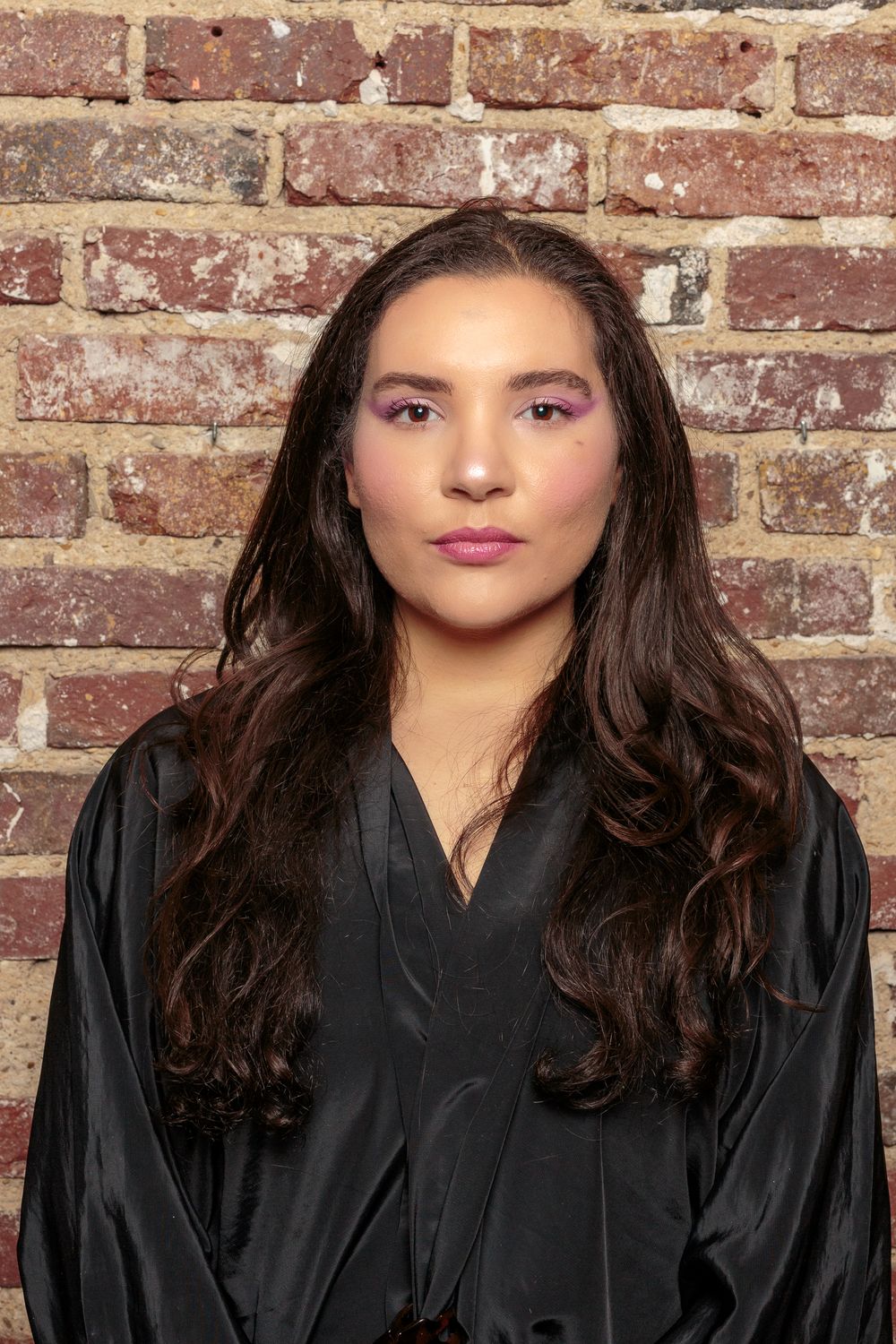 Young woman with long dark wavy hair and bold purple eye makeup posing against a brick wall.