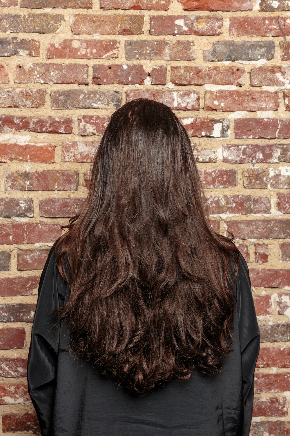 Back view of a woman with long dark voluminous wavy hair standing against a brick wall.