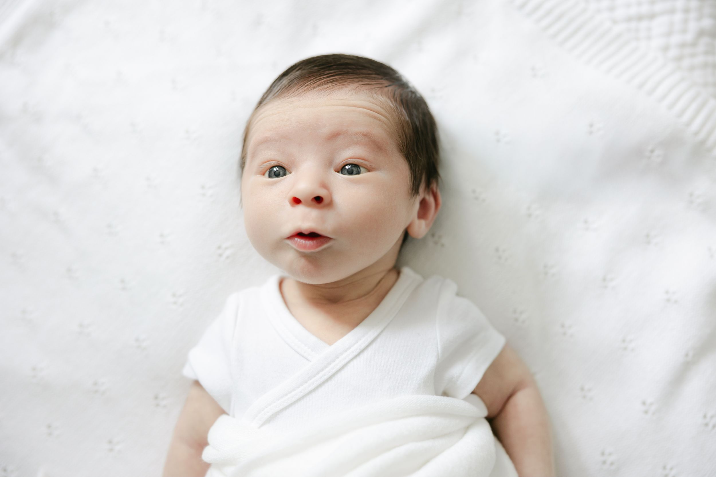 A baby wearing a white onesie lies on white bedding while looking up with a curious expression.