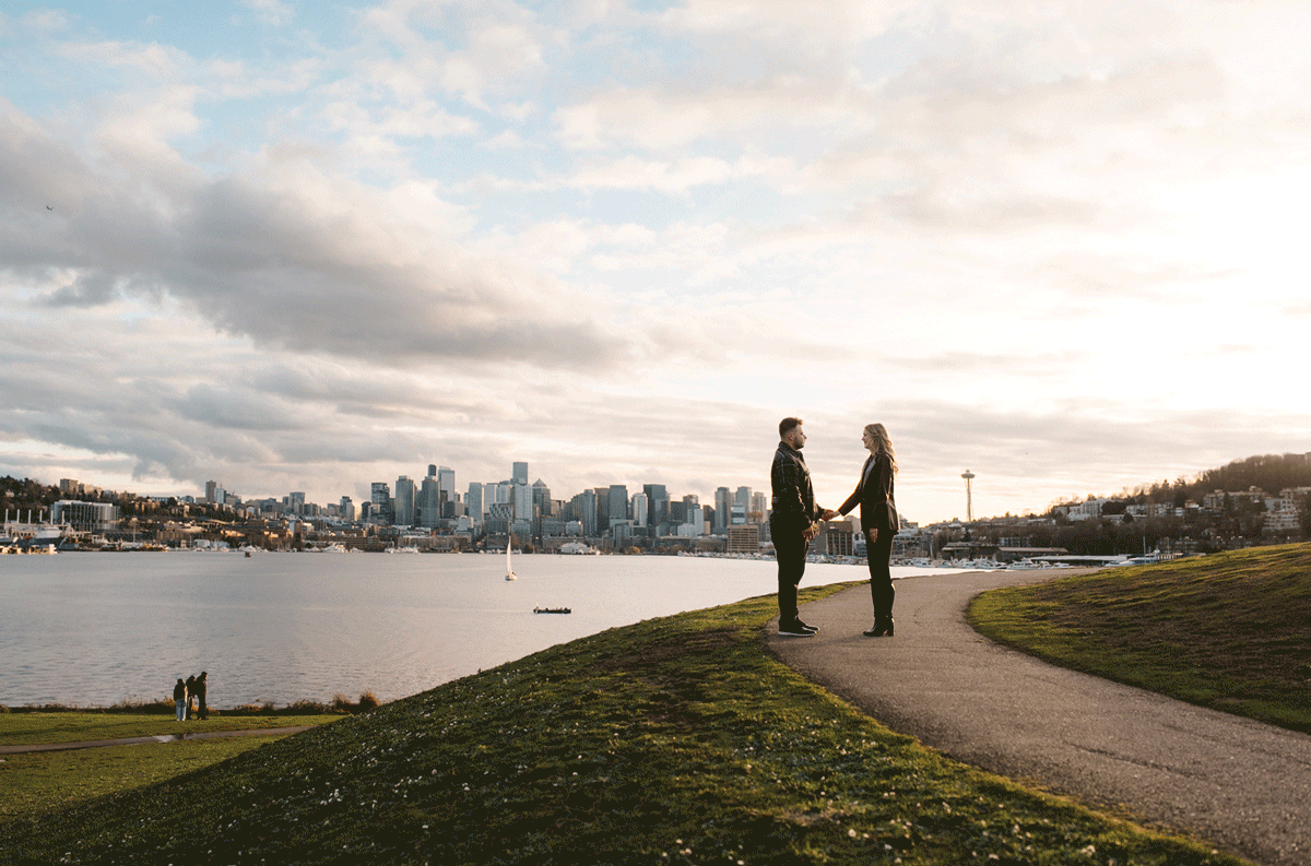 People silhouetted against a cloudy sky walk along a hilltop path with a city skyline visible in the distance.