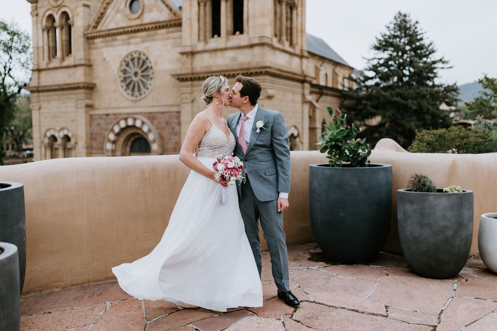 A romantic couple in wedding attire embrace outside a historic Spanish-style cathedral with adobe walls and gothic architecture.