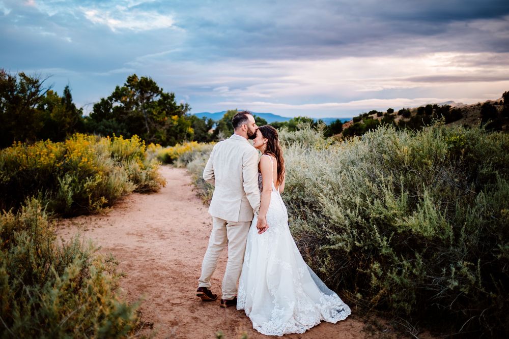A couple shares a romantic moment on a desert trail surrounded by sagebrush at sunset with dramatic cloudy skies.