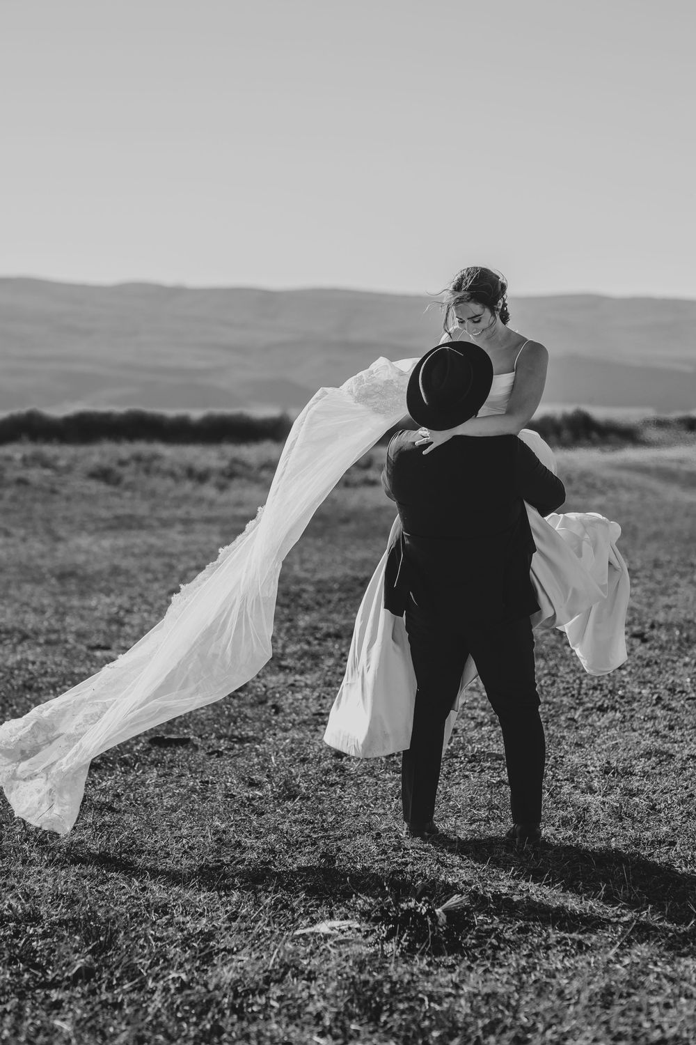 Black and white wedding photo with bride and groom embracing on hillside as veil flows dramatically in the wind.