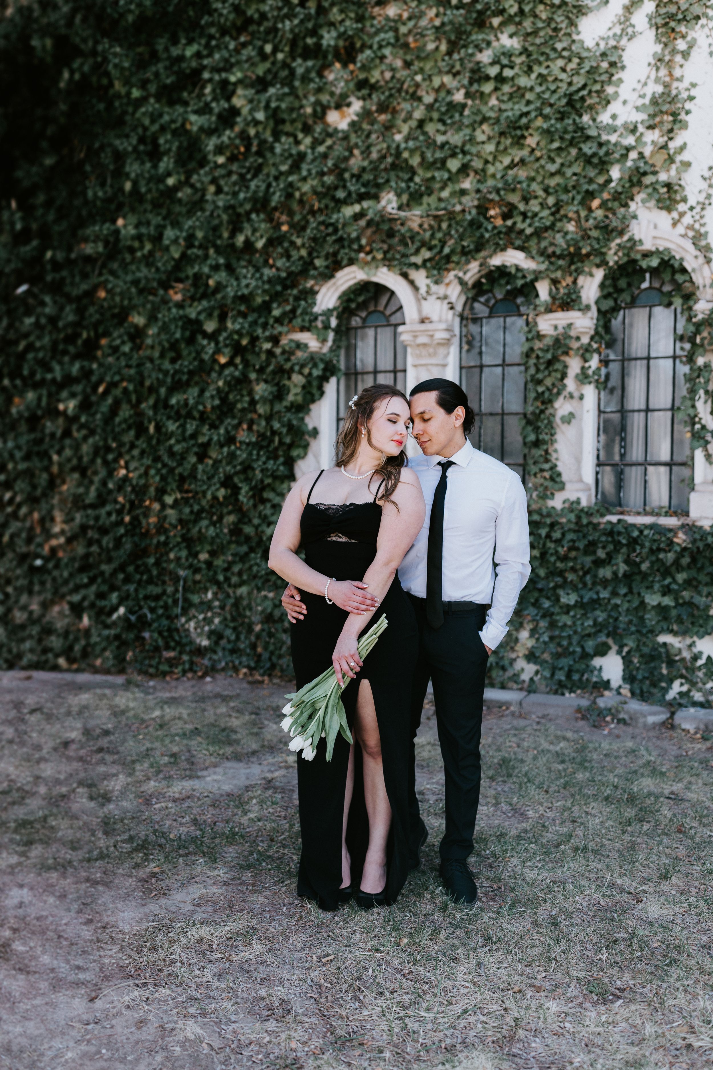 A romantic couple poses in formal attire against an ivy-covered building with arched windows in an elegant outdoor setting.