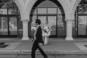 A blurred figure walks past classical stone archways while another person stands in a white outfit against the columns.