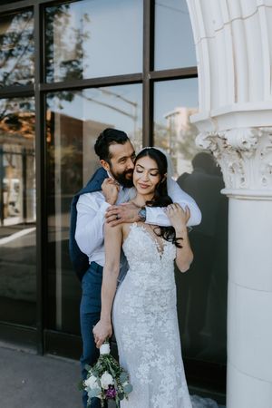 A couple shares a tender moment against an architectural backdrop, with the bride holding a floral bouquet near a white column.