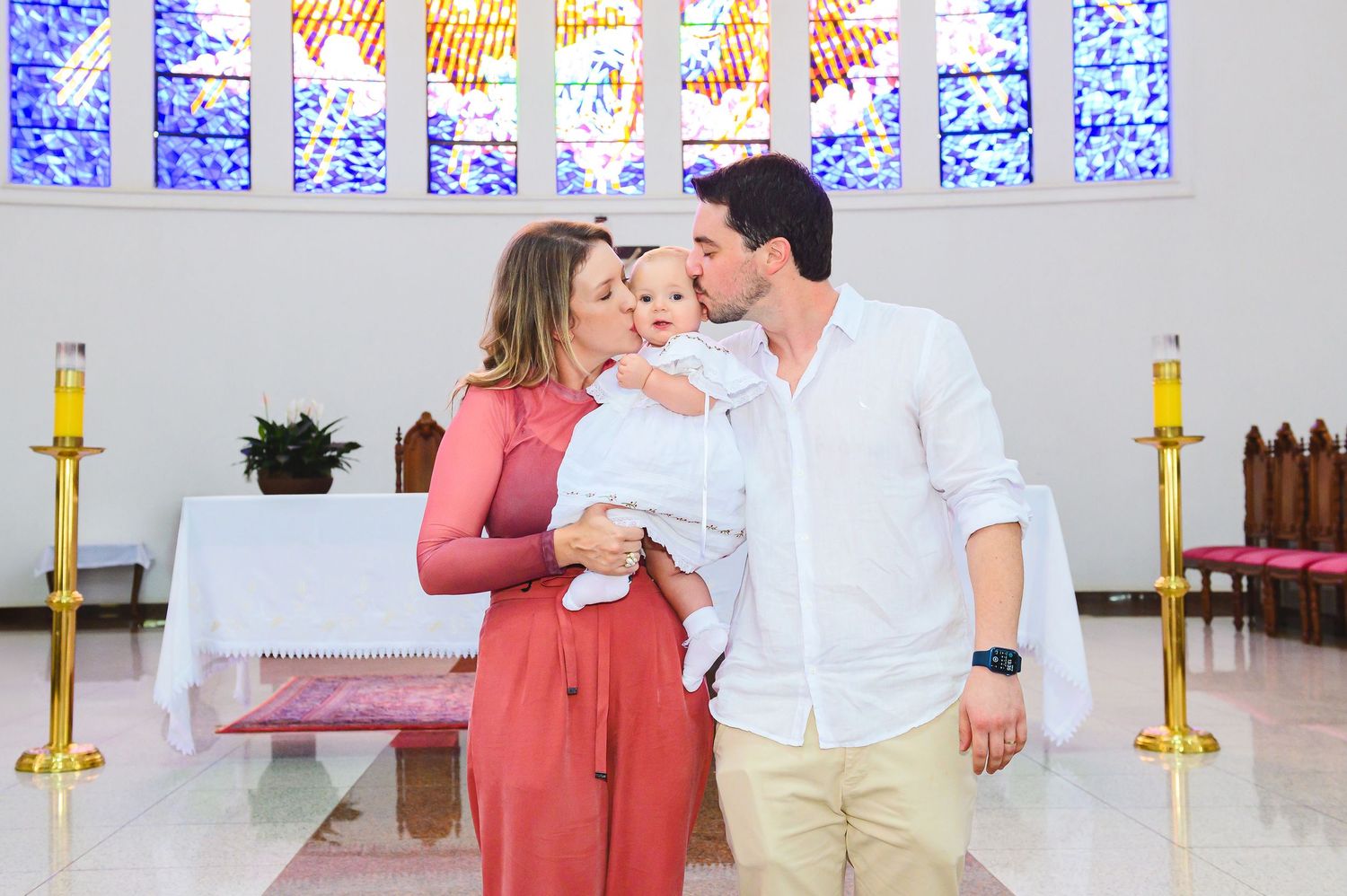 A couple shares an intimate moment inside a church with their baby, featuring stained glass windows and candle stands.