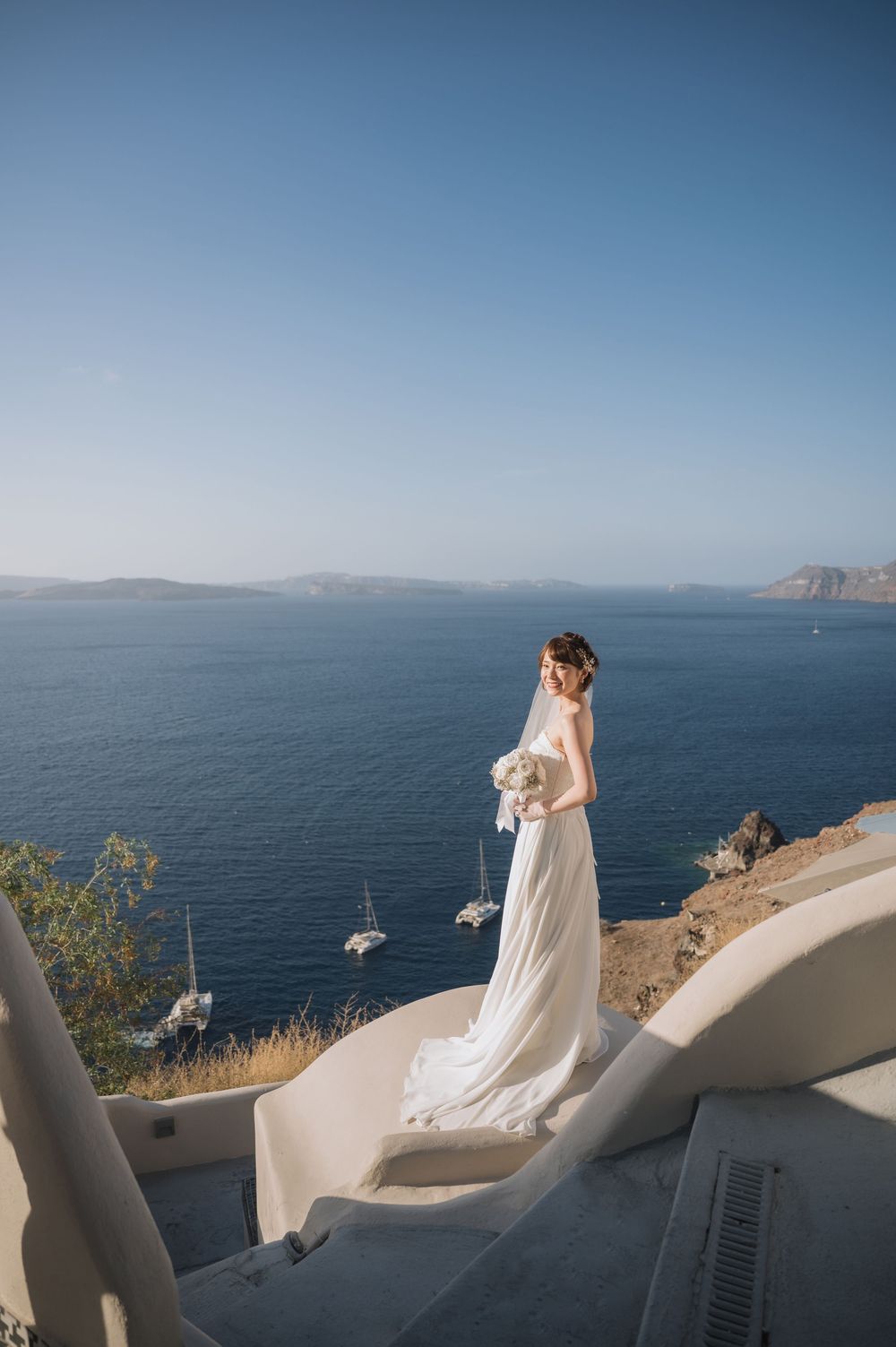 A bride in white dress stands on white steps overlooking the Aegean Sea in Santorini.