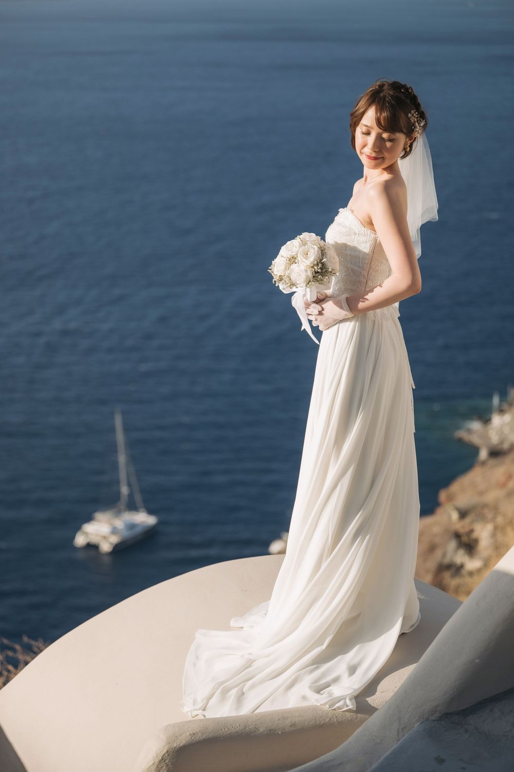 A bride in flowing white dress poses on curved white architecture overlooking blue ocean and yacht.