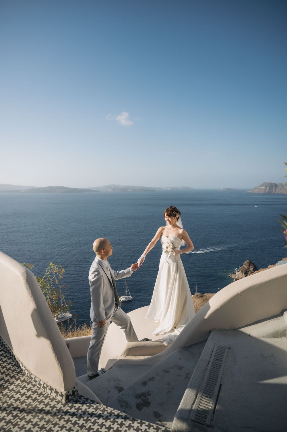 A wedding couple shares a moment on white steps with panoramic Mediterranean views.