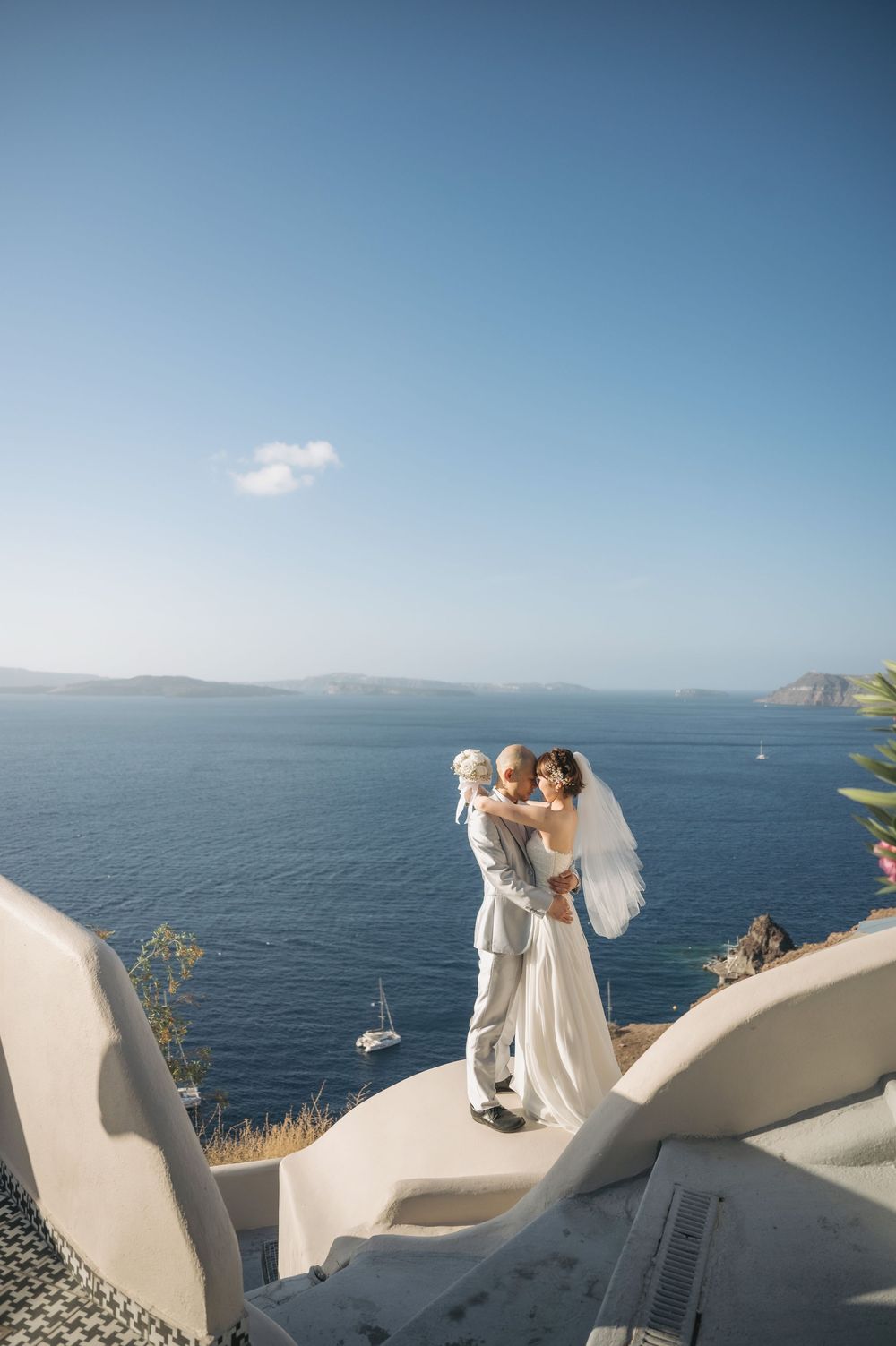 A wedding couple embraces on a white curved terrace overlooking the Mediterranean Sea with a boat in the distance.