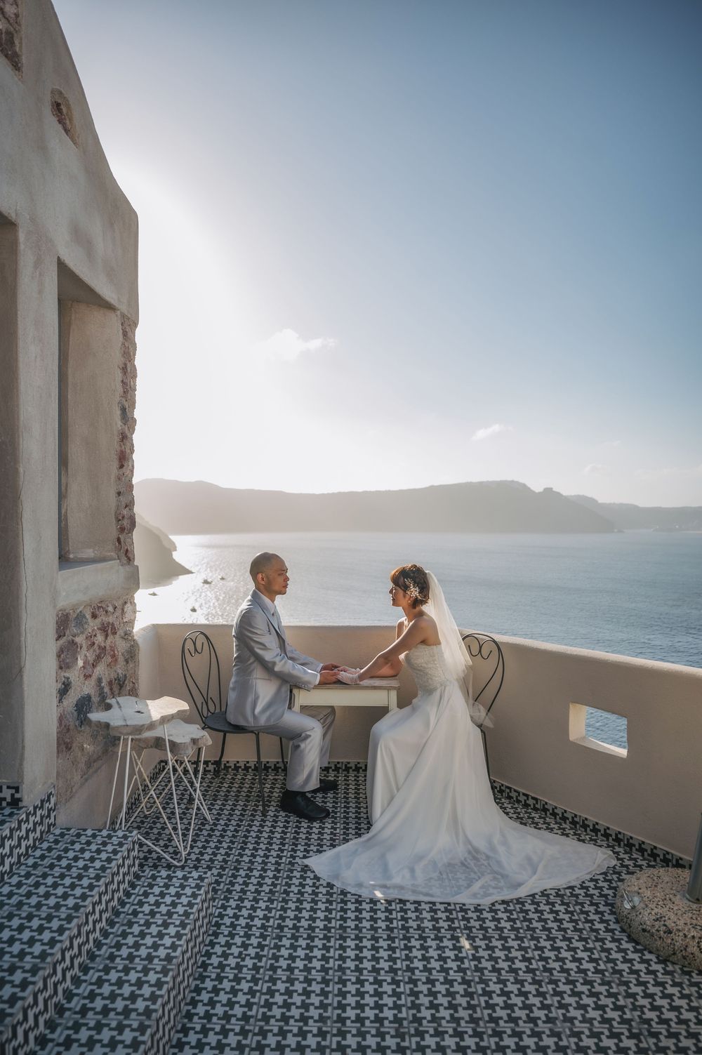 A wedding couple seated on a balcony with stunning ocean and mountain views in Santorini, Greece.