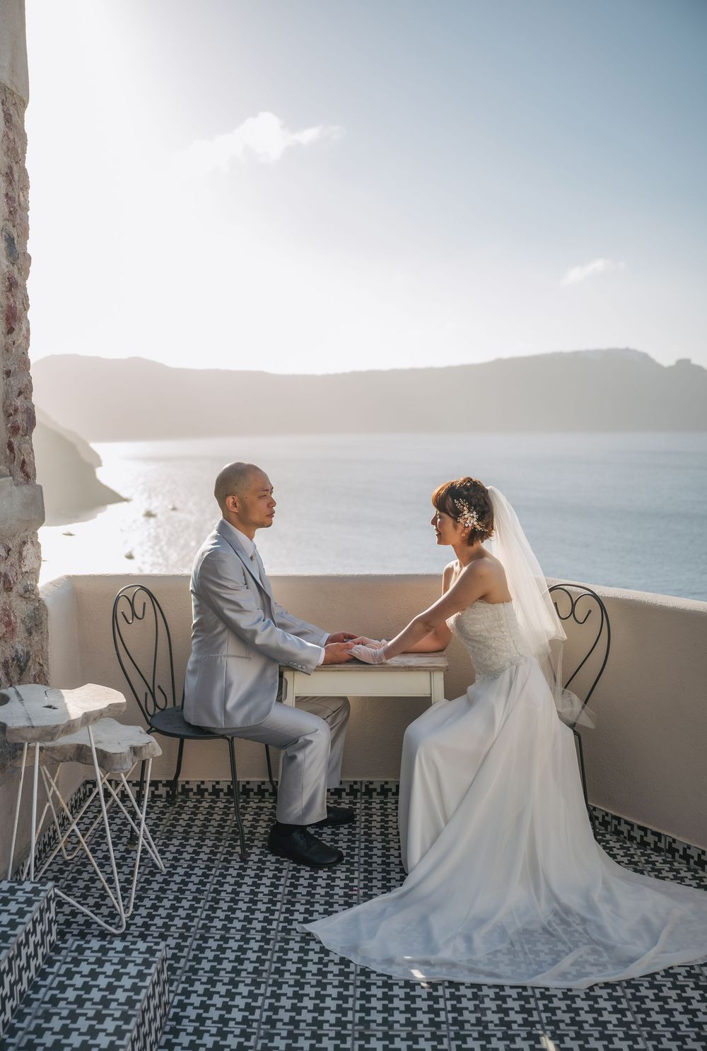 A romantic wedding couple sits on a balcony overlooking the Mediterranean Sea at sunset in Santorini.