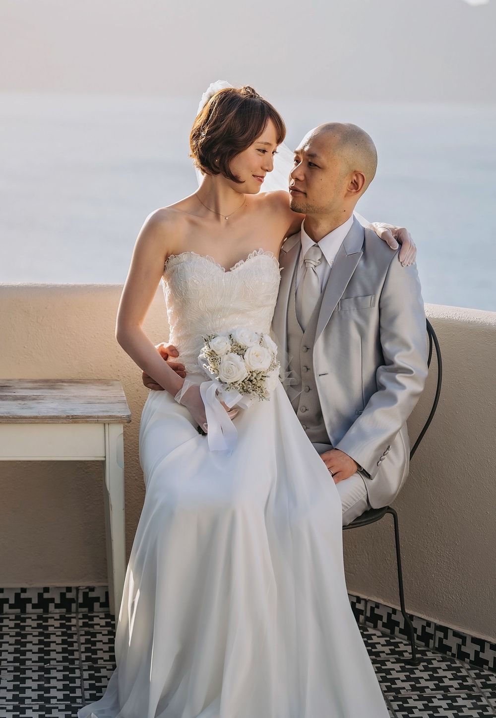 A romantic wedding couple in a strapless white gown and light gray suit pose intimately on a balcony.