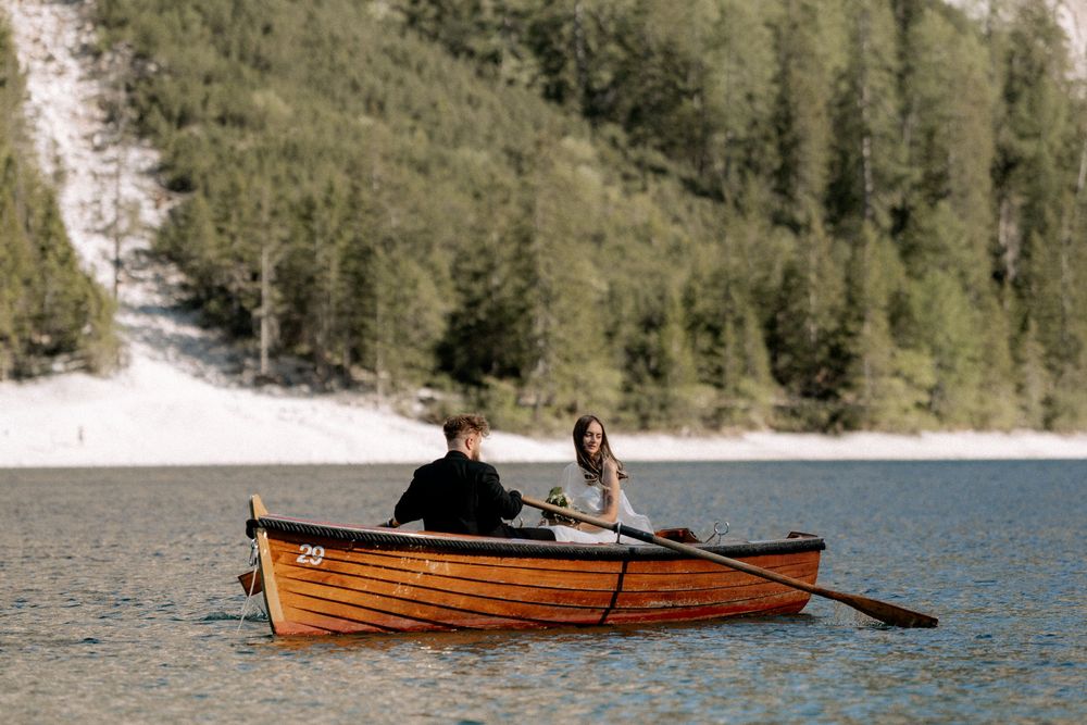Couple rowing a wooden boat on a serene alpine lake surrounded by lush green forest and snowy mountain slopes.
