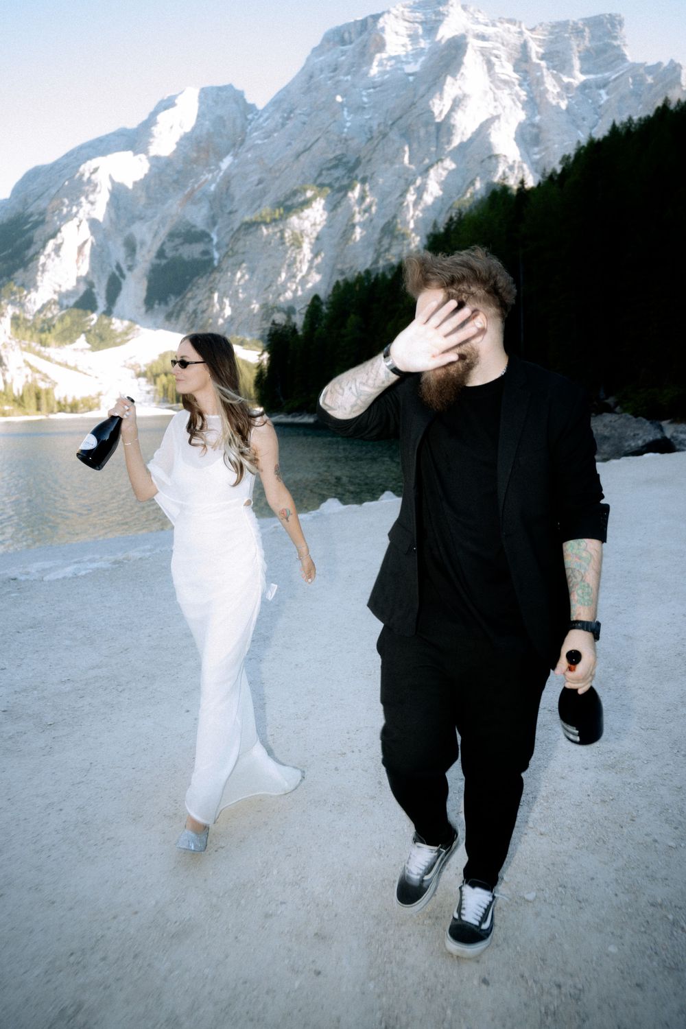 Tattooed groom in black suit and bride in white dress walk by a mountain lake during their elopement.