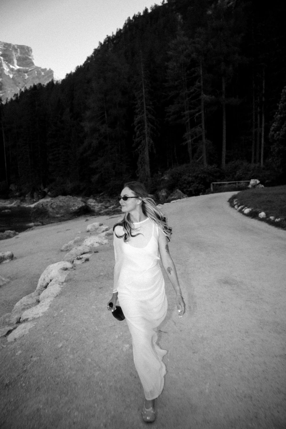 Woman in white dress walking on mountain path surrounded by tall trees, black and white photography.