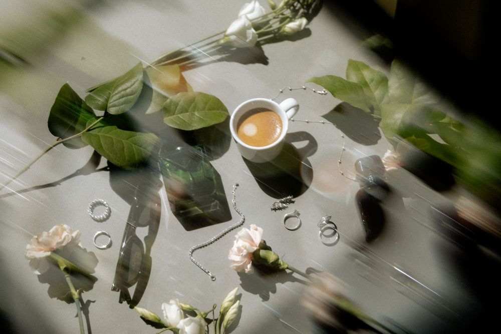 A flat lay of a coffee cup, pink roses, green leaves, and delicate rings arranged on a gray surface.